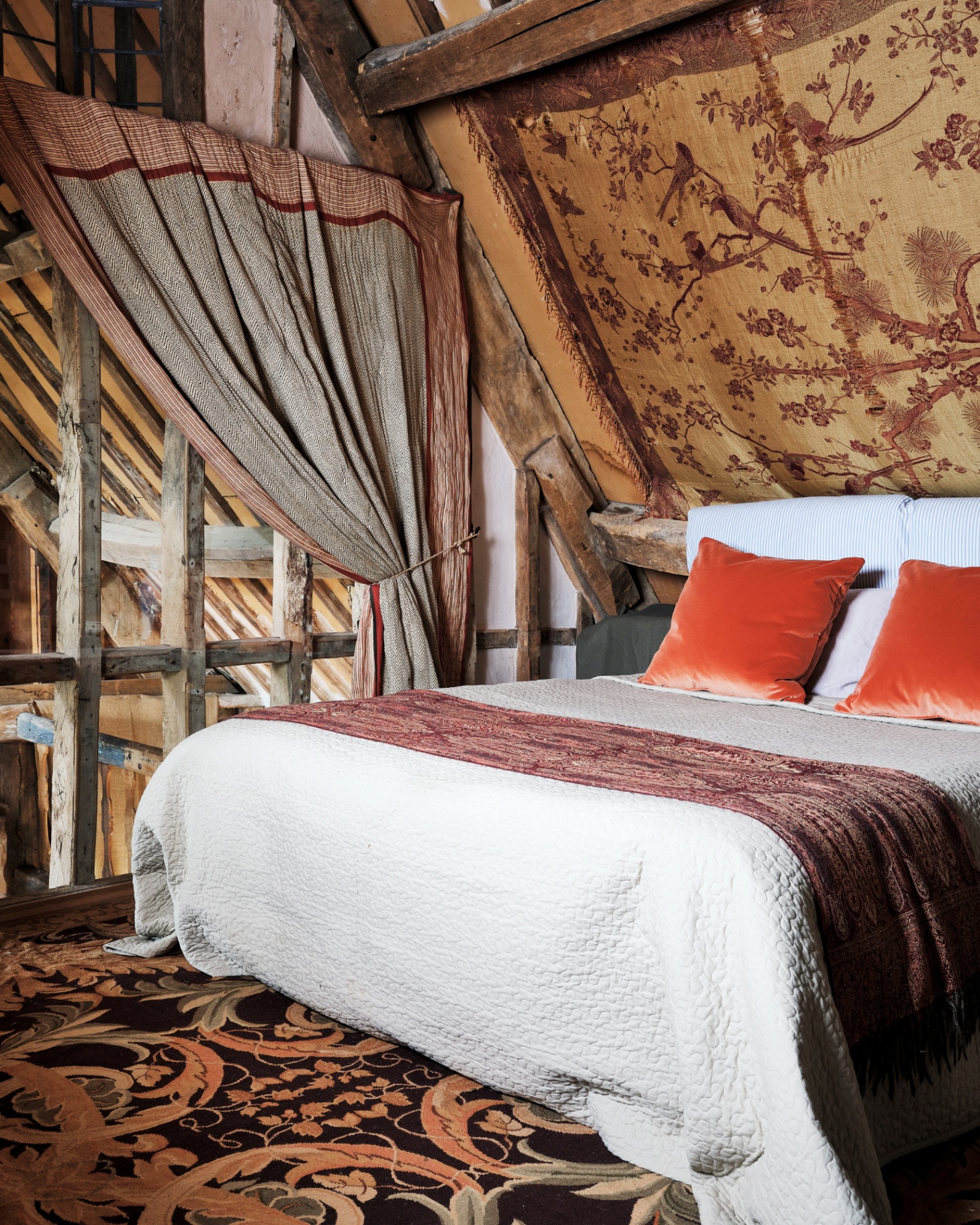 a bedroom with sloping ceilings and wooden beams, the ceiling is covered in an old tapistry style hanging, the bed has white bedding with orange velvet cushions and the floor is covered in a black and orange botanical patterned carpet