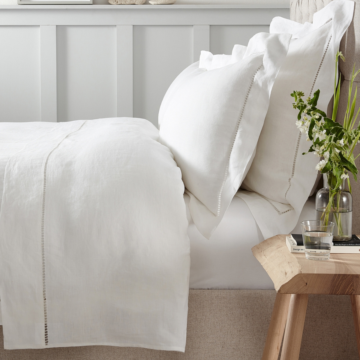 a bed with a white linen duvet cover set and a wooden bedside table with a clear glass vase and green and white flowers