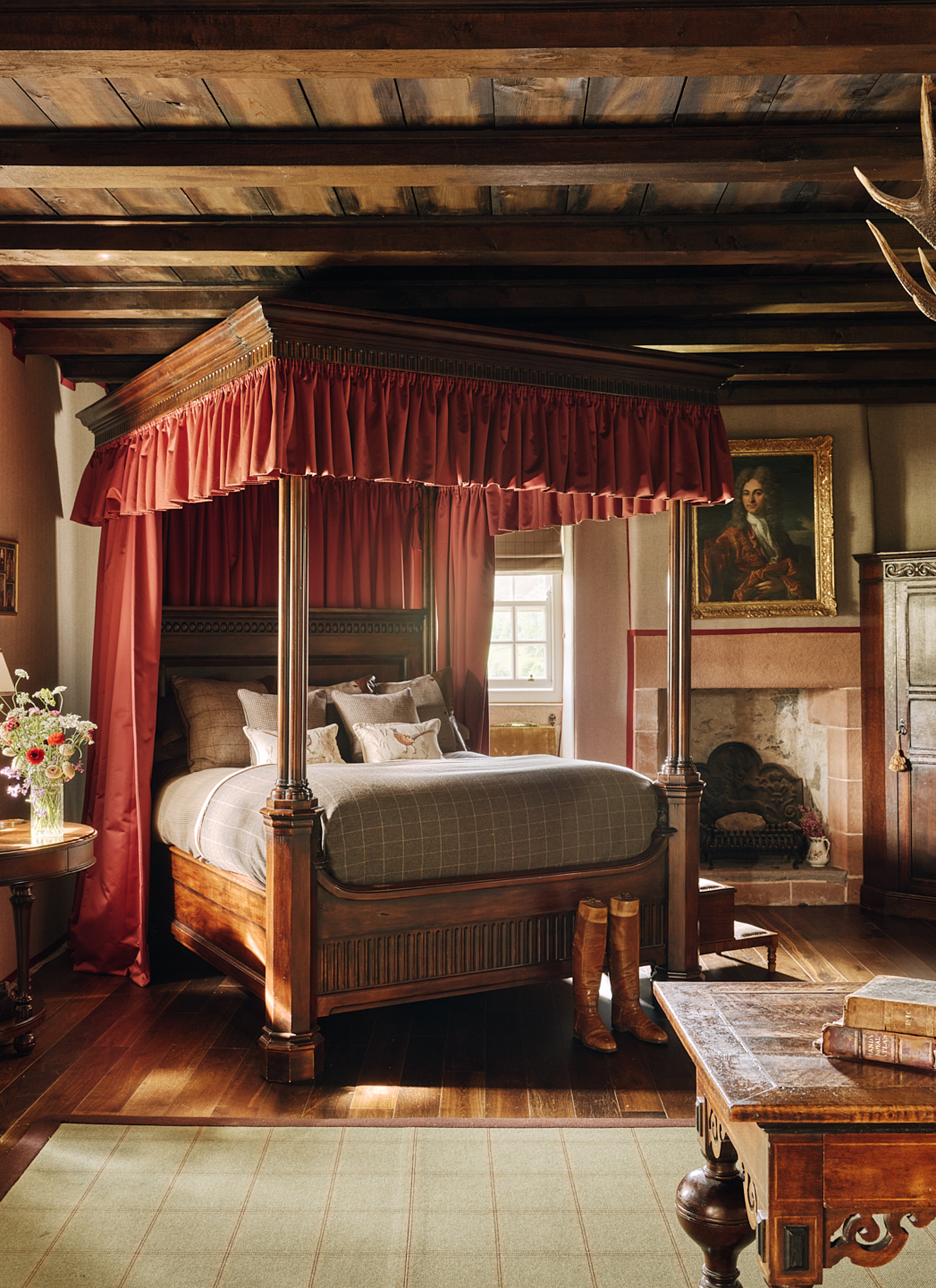 A stately, rustic bedroom centered around a heavy dark timber four-poster bed draped in deep red fabric, set beneath exposed wooden ceiling beams with a stone fireplace, framed oil portrait, and rich hardwood flooring.