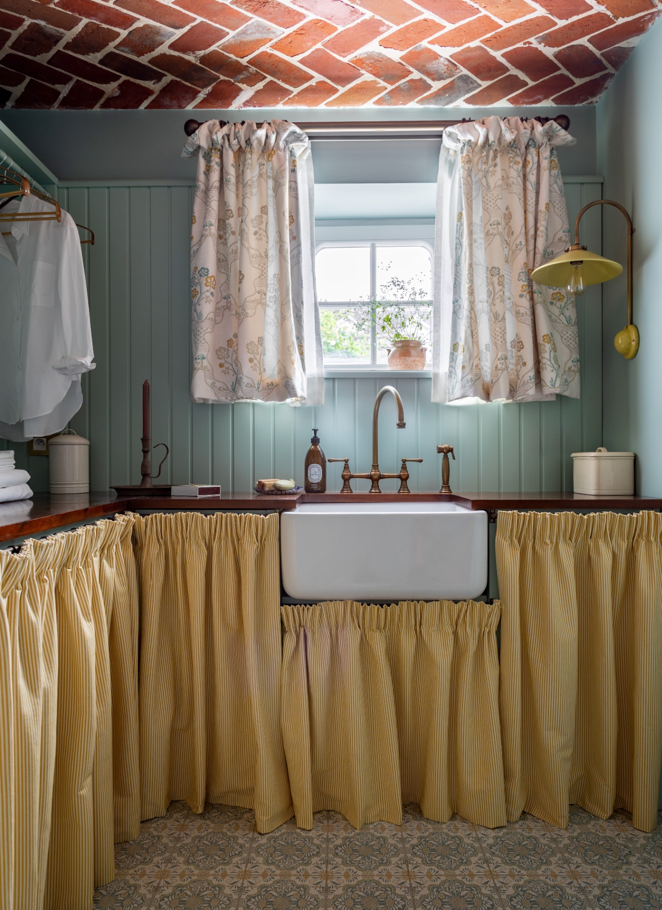 a country utility room with pale blue walls, a yellow ad white stripe curtain under the counter tops, a belfast sink and a window with white floral curtains