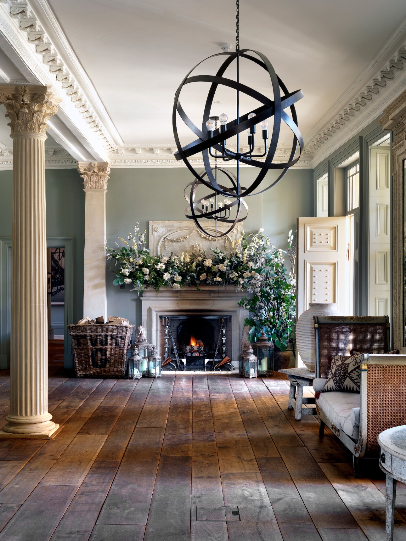 Jade Holland Cooper’s grand entrance hall with classical stone columns, a lit fireplace, floral mantle display, and large orb chandeliers