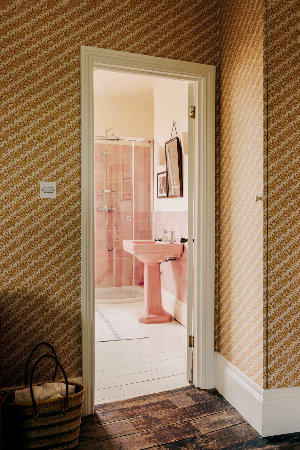 In the foreground is a wall covered in a diagonal yellow small-scale wallpaper. Through the open doorway a bathroom can be seen with a pink basin and pink-tiled shower