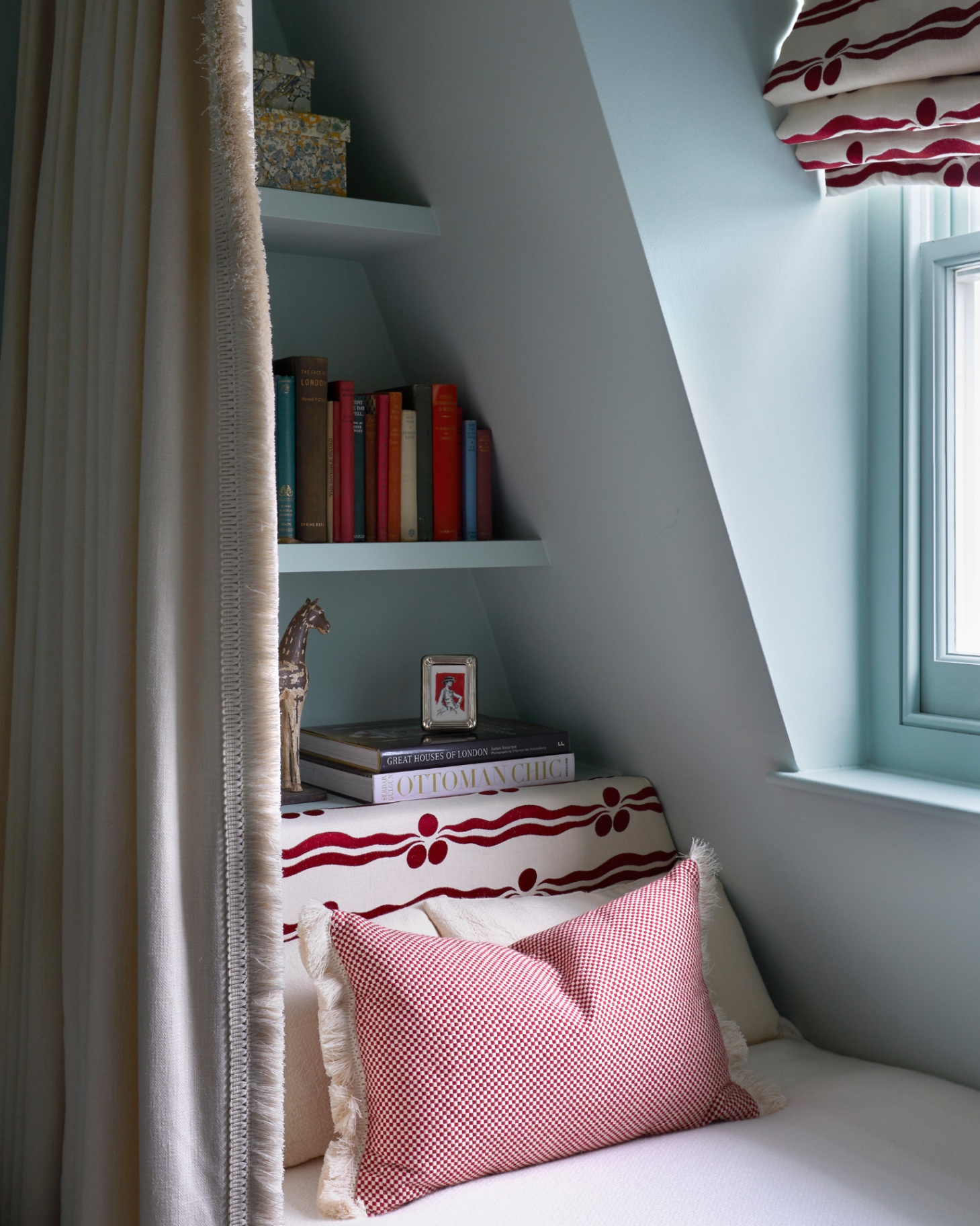 a bed nook with blue painted recessed shelving above the headboard filled with books and toys and a single bed with a large red and white patterned cushion