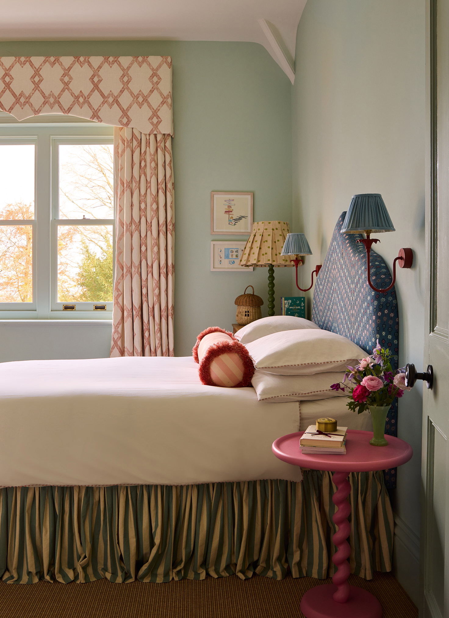 a bedroom with a bed with a striped valance and a blue patterned headboard, two red and blue pleated sconces either side and a bright pink bedside table with a vase of flowers