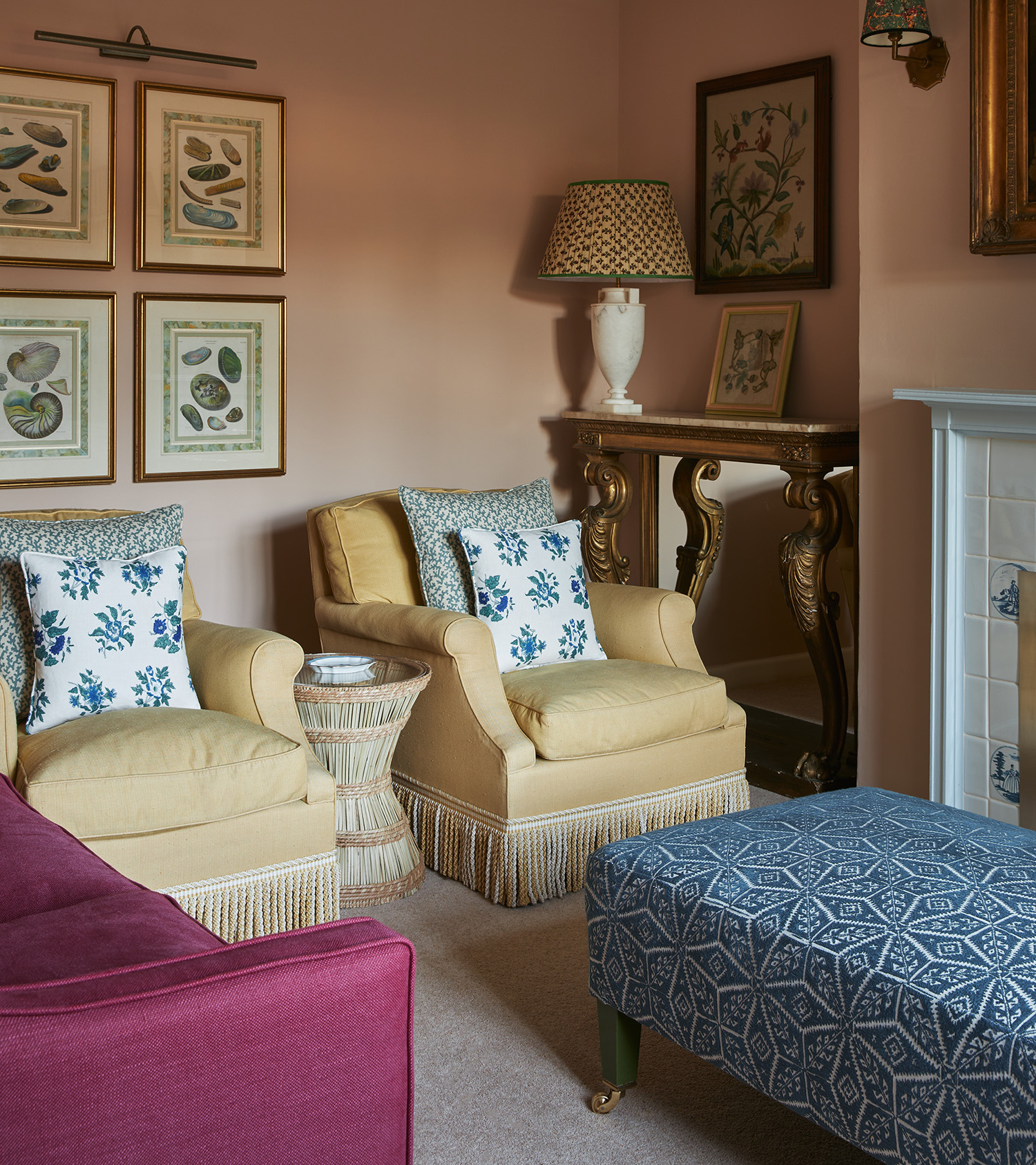 a living room corner with plaster pink walls, two pale yellow armchairs with tassels, art on the wall and a blue patterned ottoman 