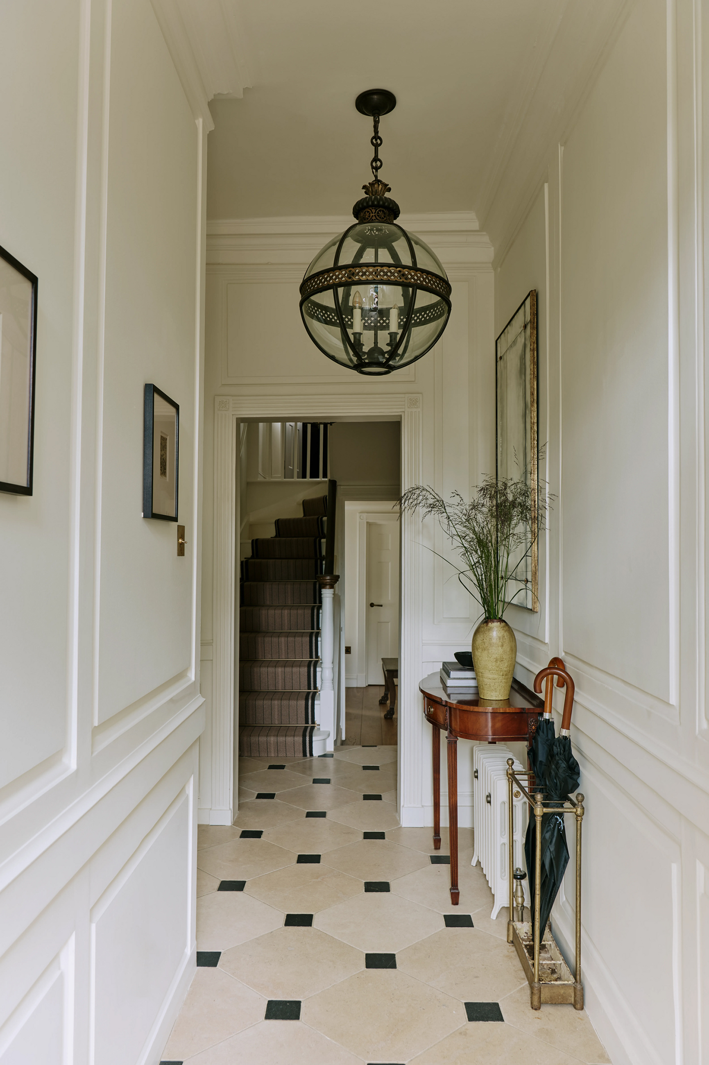 A hallway in a townhouse with tiled black and white floor, globe pendant lantern, wooden semi-circular console table and umbrella stand. In the background, stairs can be seen, decorated with a striped runner, leading upstairs 