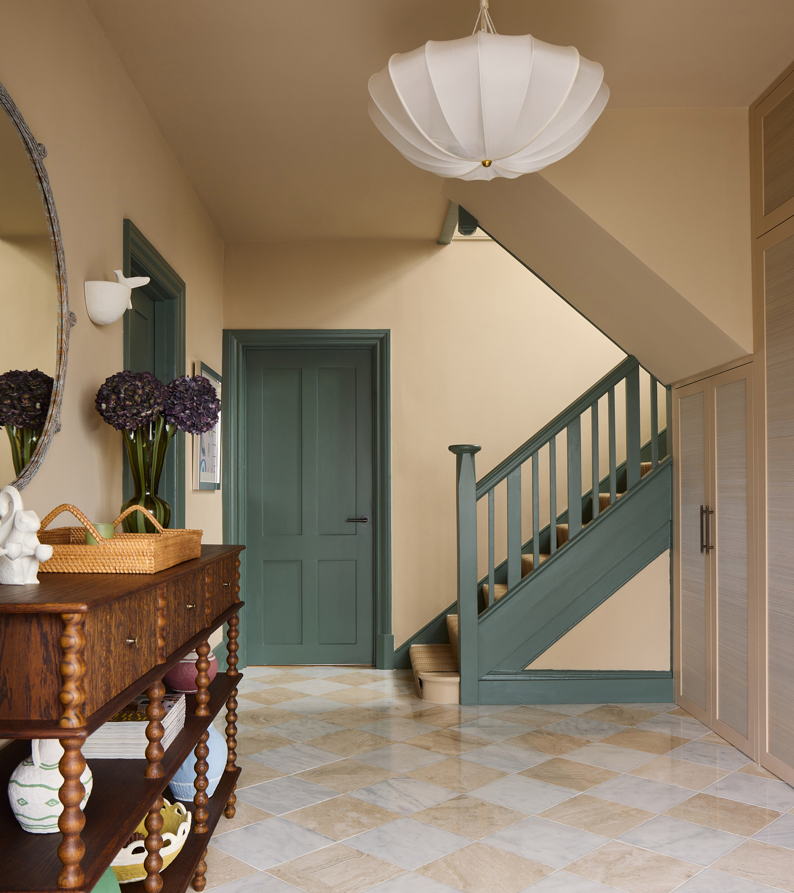 a large hallway with beige walls and teal painted doors and stair banister, beige and white checkered floors and a wooden bobbin console table