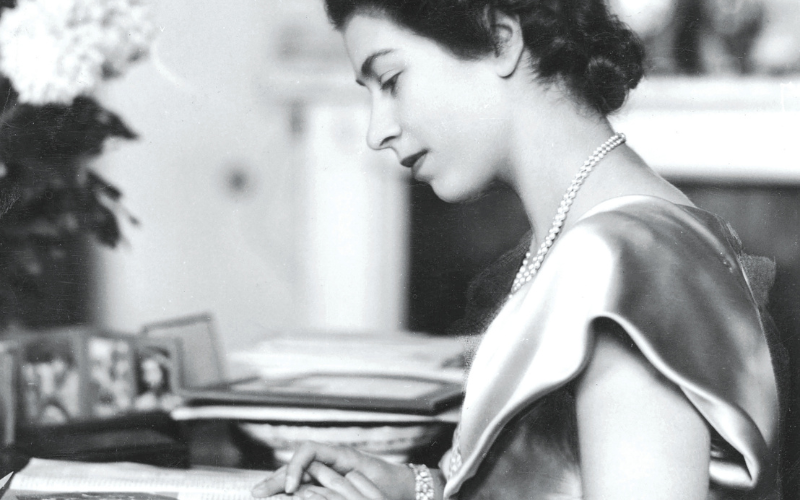 Princess Elizabeth aged 21 at Buckingham Palace, the photograph is in black and white and she wears a silk dress and a pearl necklace, she sits at a desk writing a letter