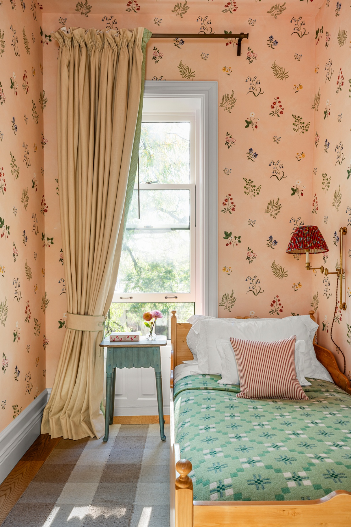 A small bedroom with a single wooden bed to the right and a dainty blue bedside table beside it, in front of a large window dressed with pleated curtain. A check rug covers floorboards and the walls and ceiling are in a pretty wildflower wallcovering