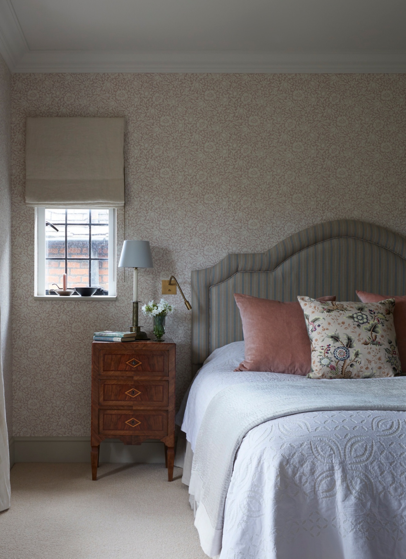 a bedroom with pale floral wallpaper, a dark brown wooden bedside table with avase of flowers and books on it and a bed with a striped headboard and white bedding