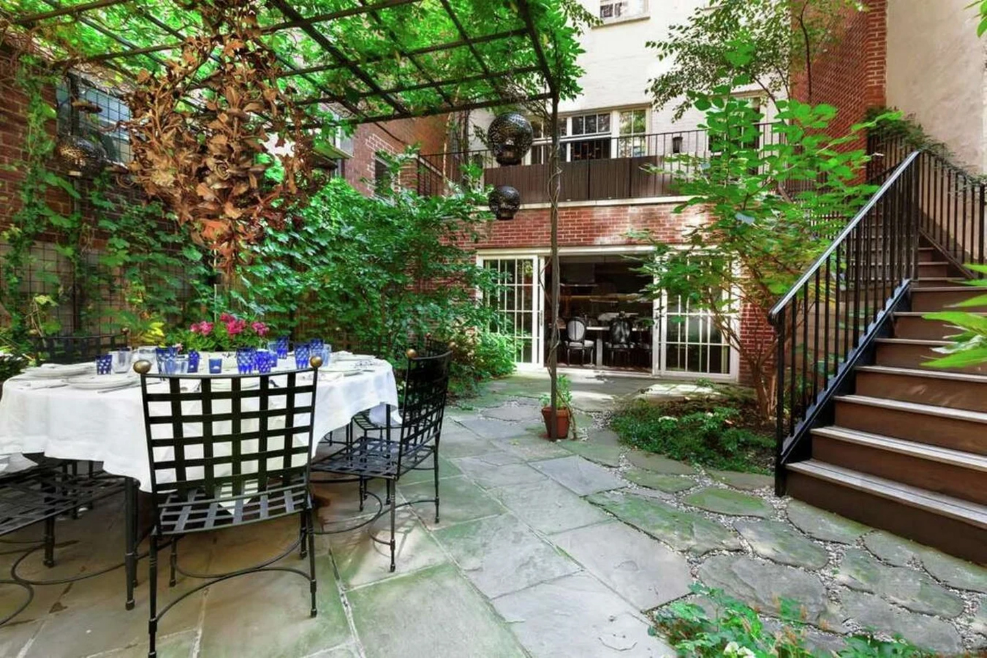 the outside of a house with a patio, red brick, a large staircase, and a pergola with greenery and a dining table and chairs underneath