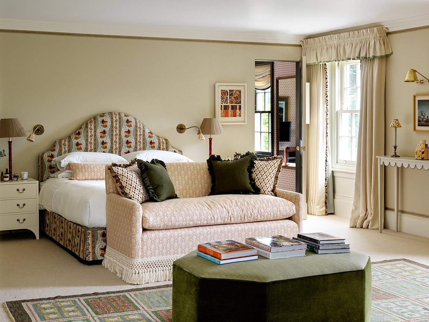 A large bedroom with a double bed upholstered in a floral stripe in the background. At the end of the bed is a fringed pink patterned sofa and a long octagonal velvet green ottoman topped with books sat on a patterned rug