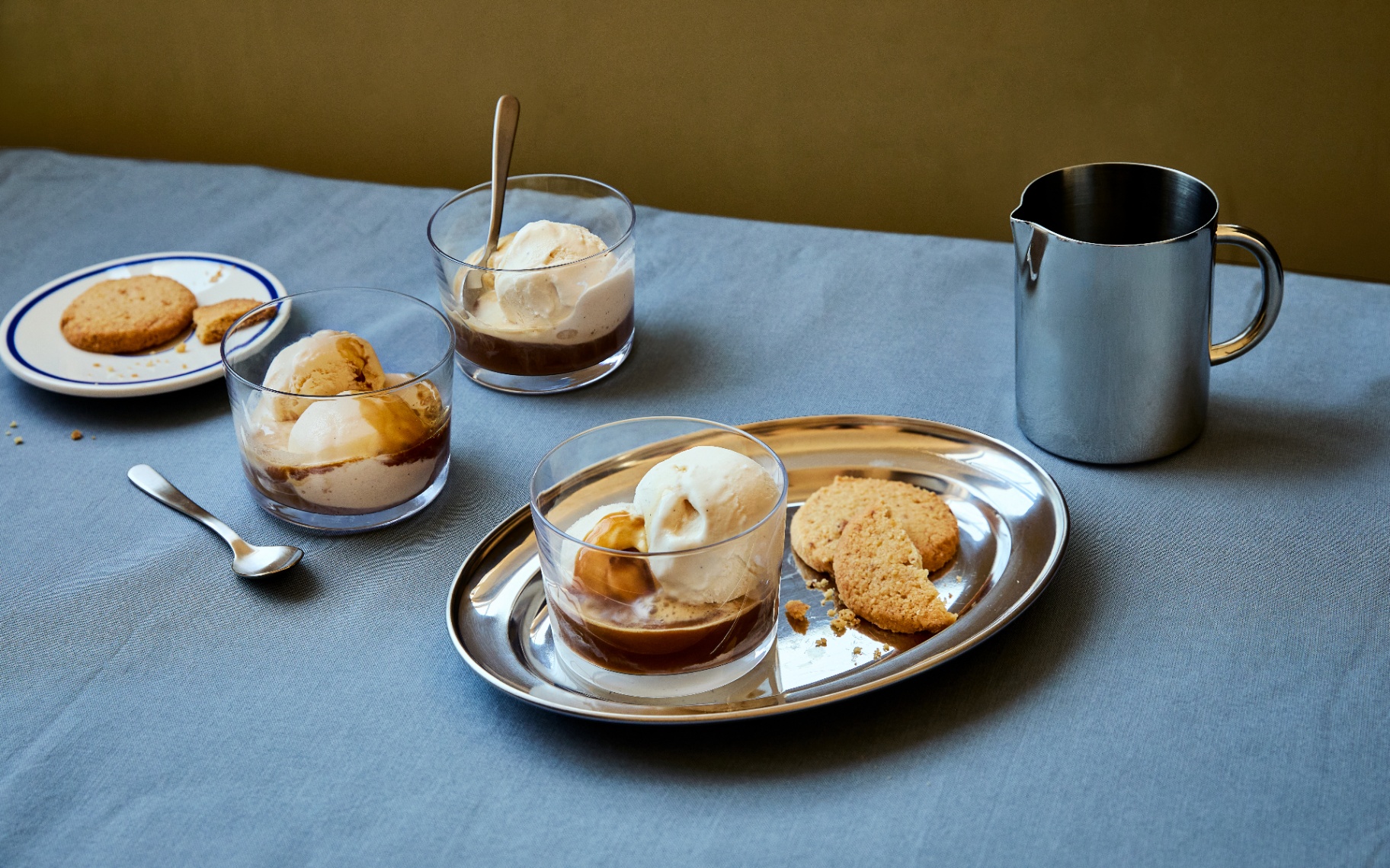 3 small clear glass bowls of affogato with a stainless steel pitcher on a blue table cloth