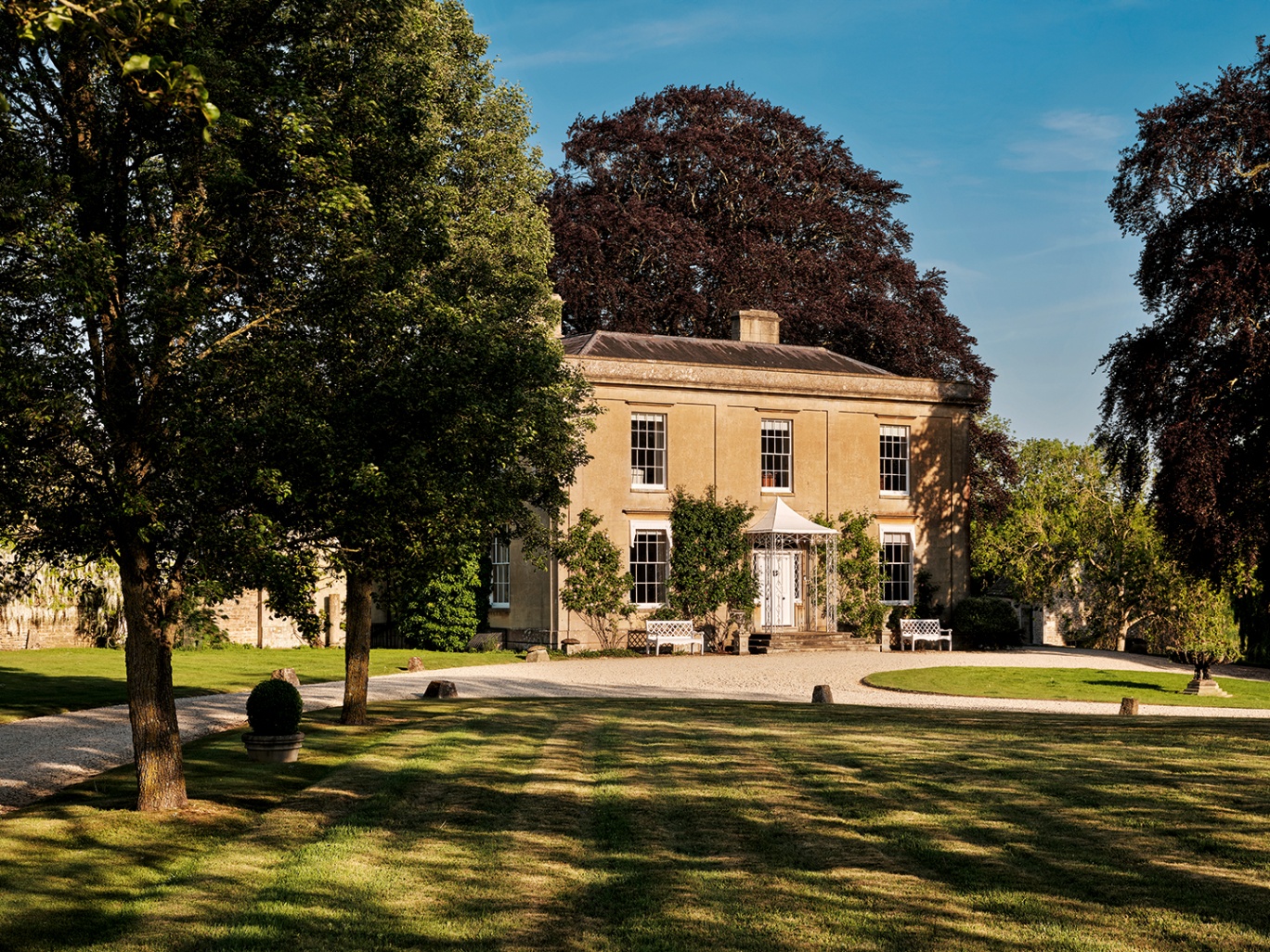 a large country manor house with five sash windows and a front door with a canopy above it, surrounded and half covered by large trees
