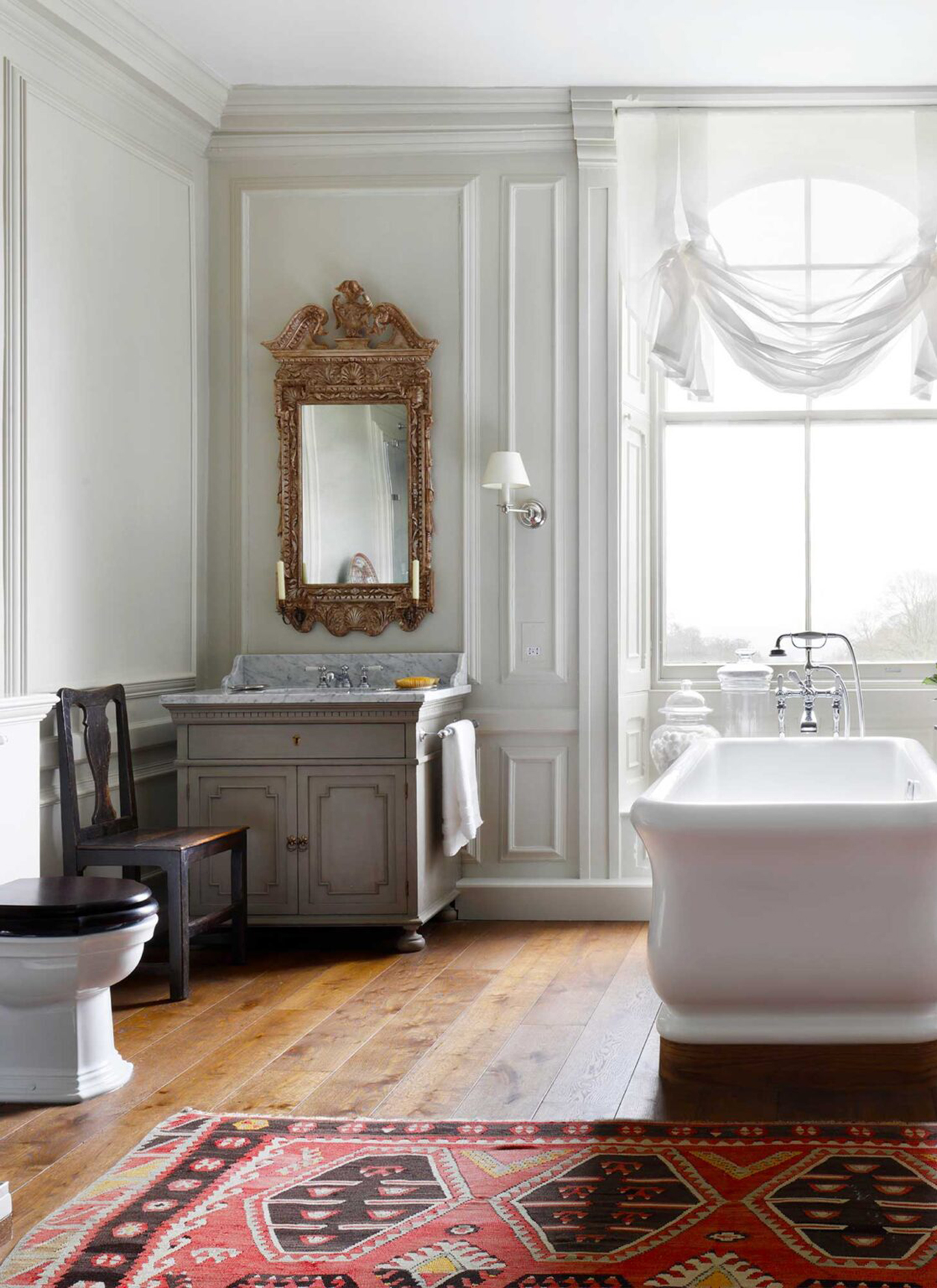 a bathroom with white panelled walls, a freestanding bath tub, wooden floors and a red patterned rug