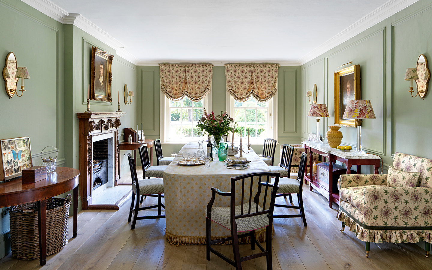 A green dining room with a large dining table, covered with a tasselled tablecloth, and upholstered dining chairs. A frilled pattern armchair and console table sits to the right, and a demi-lune console and fireplace to the left