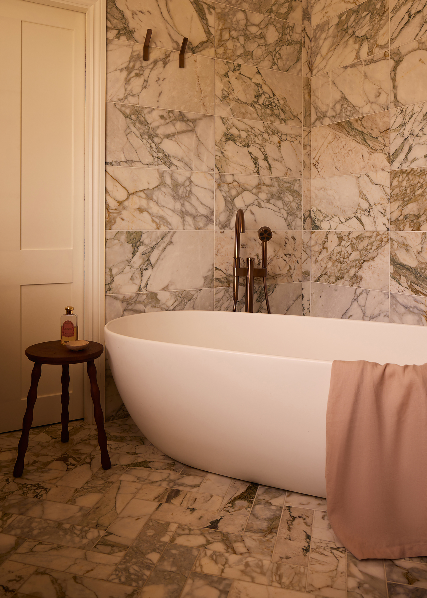 a bathroom with marble tiled floors and walls. A freestanding white bath tub is paired with a contemporary freestanding tap and has a towel draped over the side. A wooden side table sits at the side with a bottle of bath soak and soap dish on it