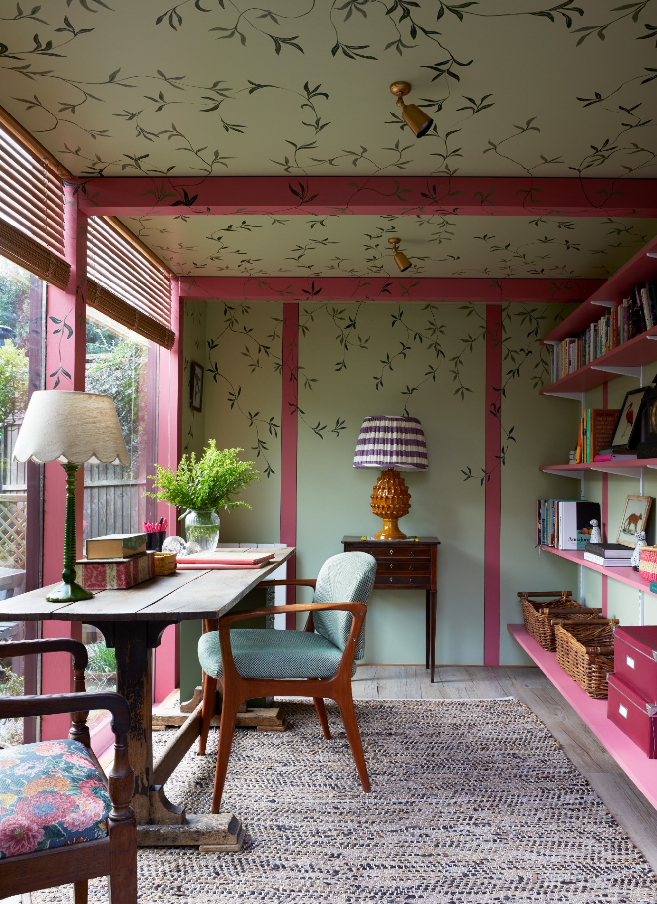 a home study with red painted beams and green walls with hand painted leaves, a desk facing the window with a blue and wooden chair and red wall shelves on the back wall