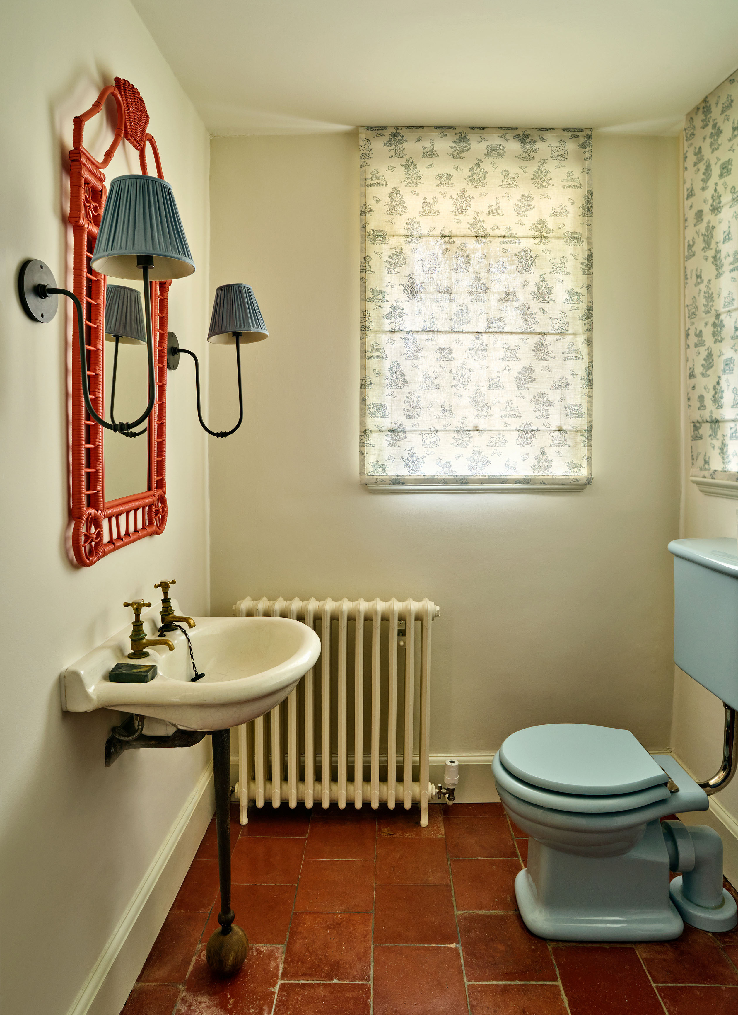a downstairs toilet with brick flooring, a pale blue toilet, a white cast iron radiator, a white pedestal sink, a coral rattan mirror and two blue pleated sconces