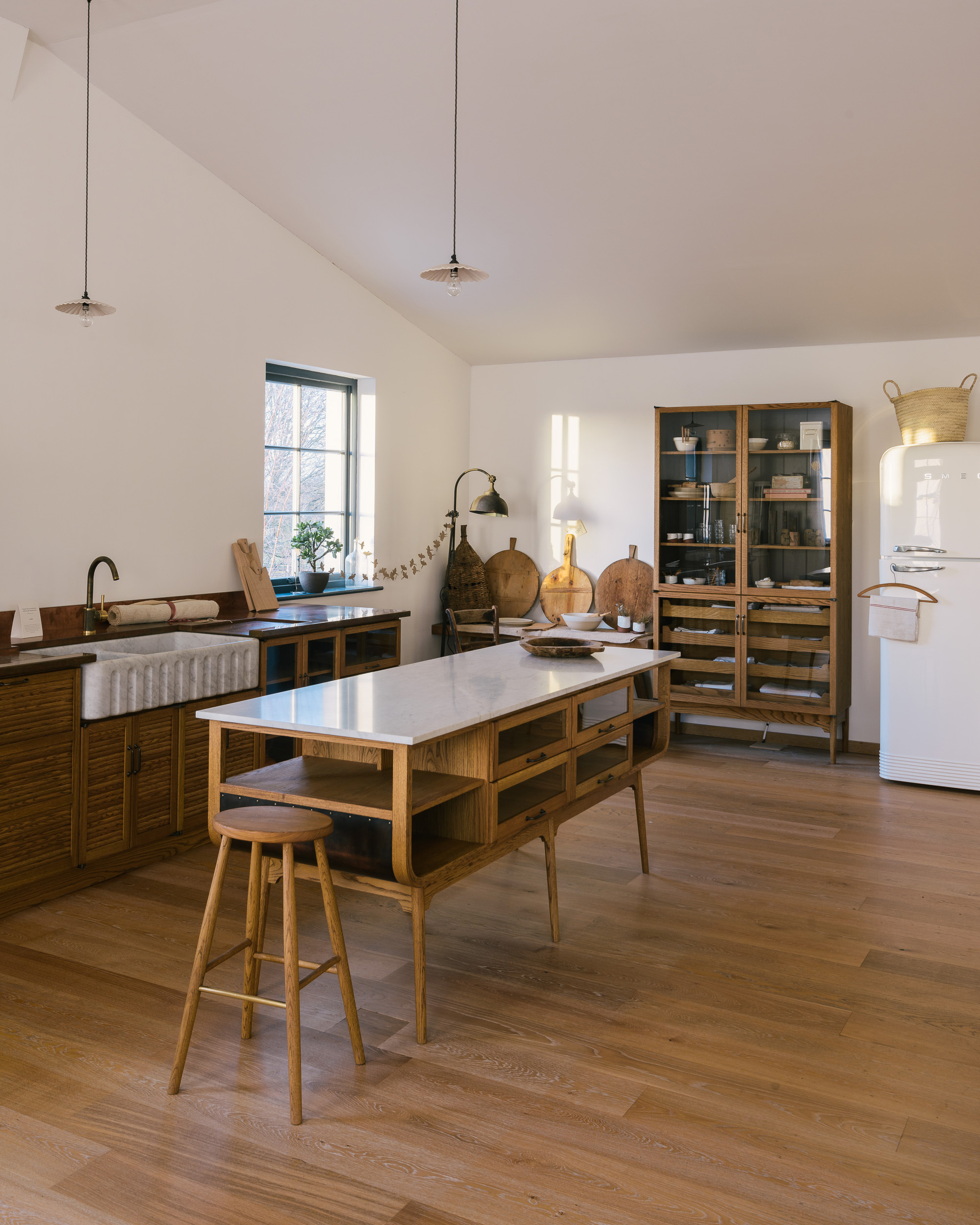 A kitchen with wooen floors, a mid century style wooden kitchen island and stalls, a white smeg fridge and panelled wooden cupboard doors