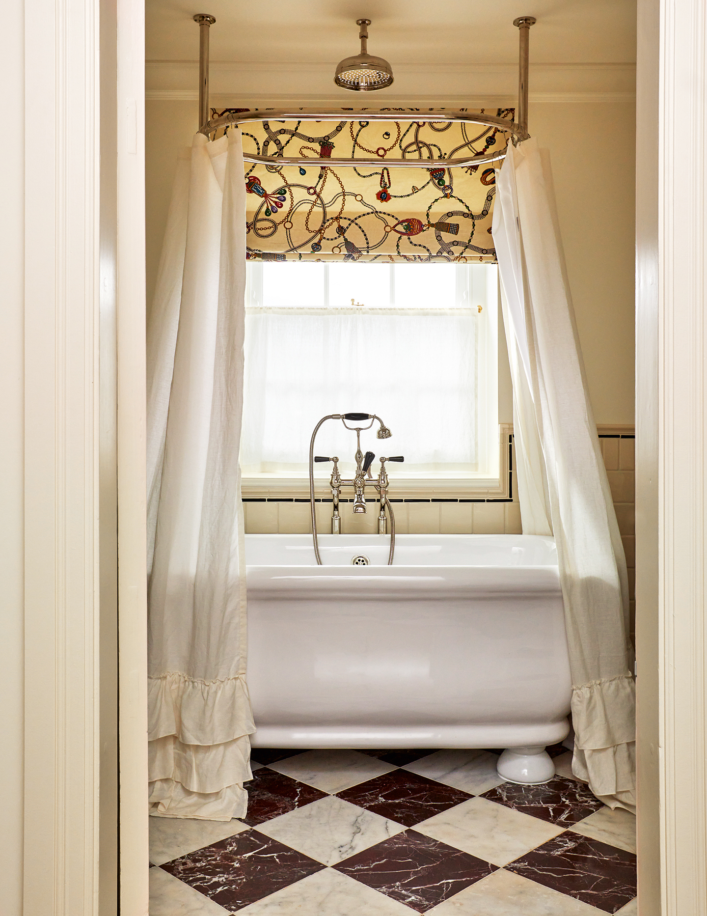 A bathroom with a checkerboard marble floor and large white freestanding bath paired with frilled shower curtains hanging off an oval rail. The lower half of the walls are tiled. The window is dressed with a cafe curtain and vintage fabric blind