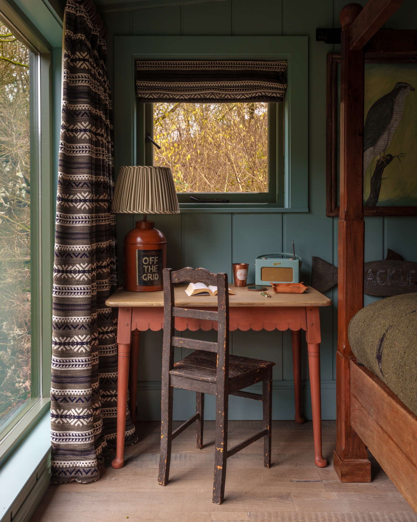 with an orange scalloped bedside dressing table with a brown painted chair and a large ceramic lamp on the table with the words off the grid. An aztec pattern set of curtains on the window to the left and a dark wood bed to the right