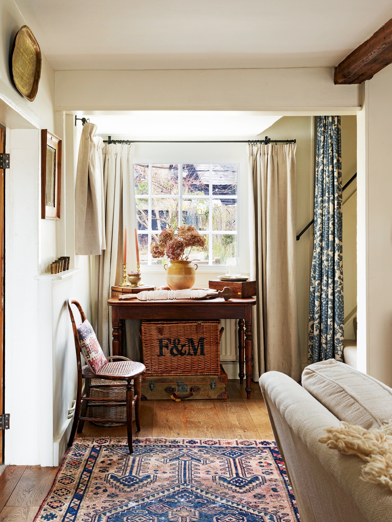 A console table with a F&M basket underneath sits in front of a window dressed with neutral curtains. On the table sits some flowers in a vase and collected objects. In front is a small cane chair and traditional patterned rug