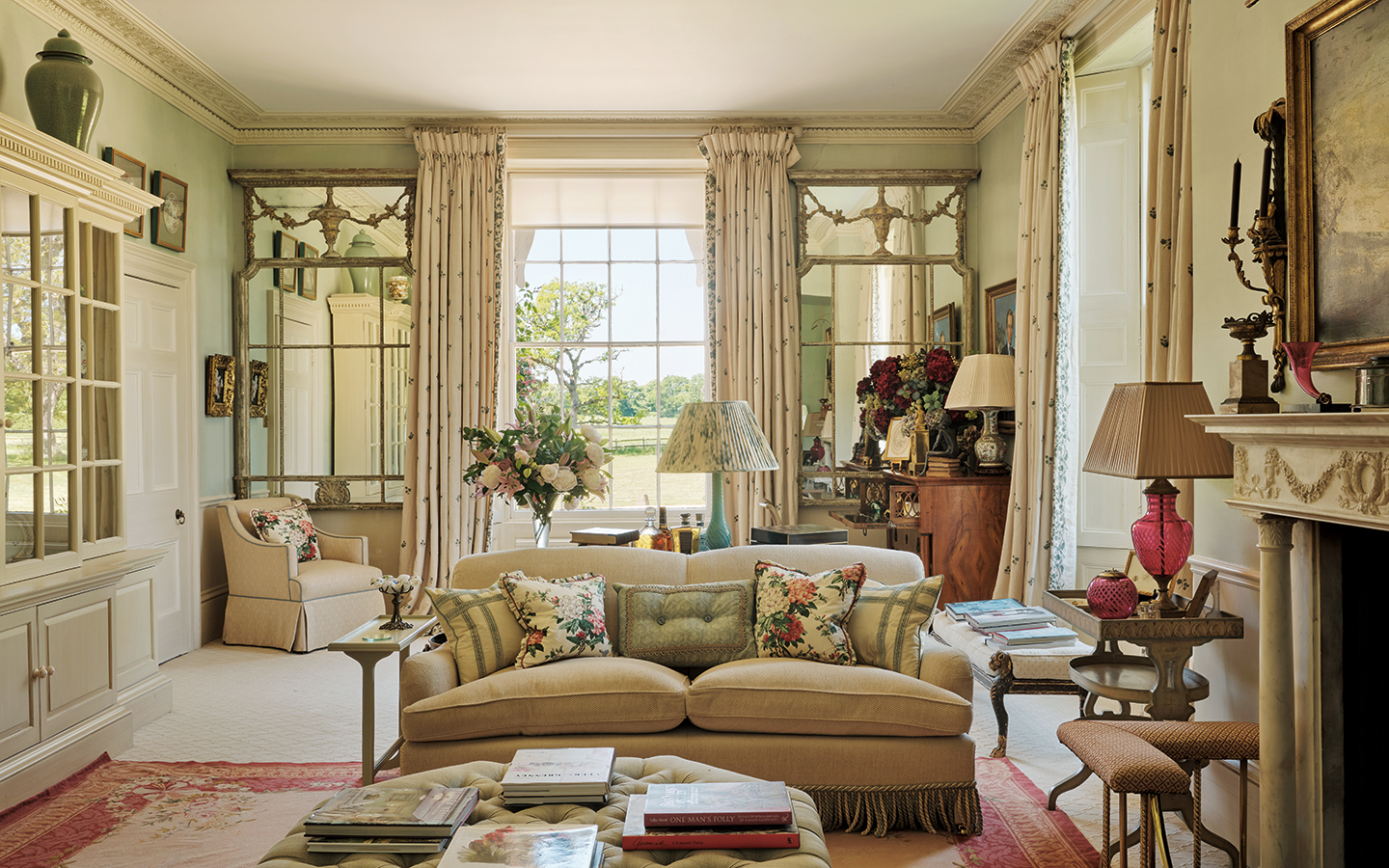 an elegant blue living room with an ottoman in the foreground, a cream sofa behind and in the background is a large sash window flanked by two large ornate mirrors and tall floral curtains