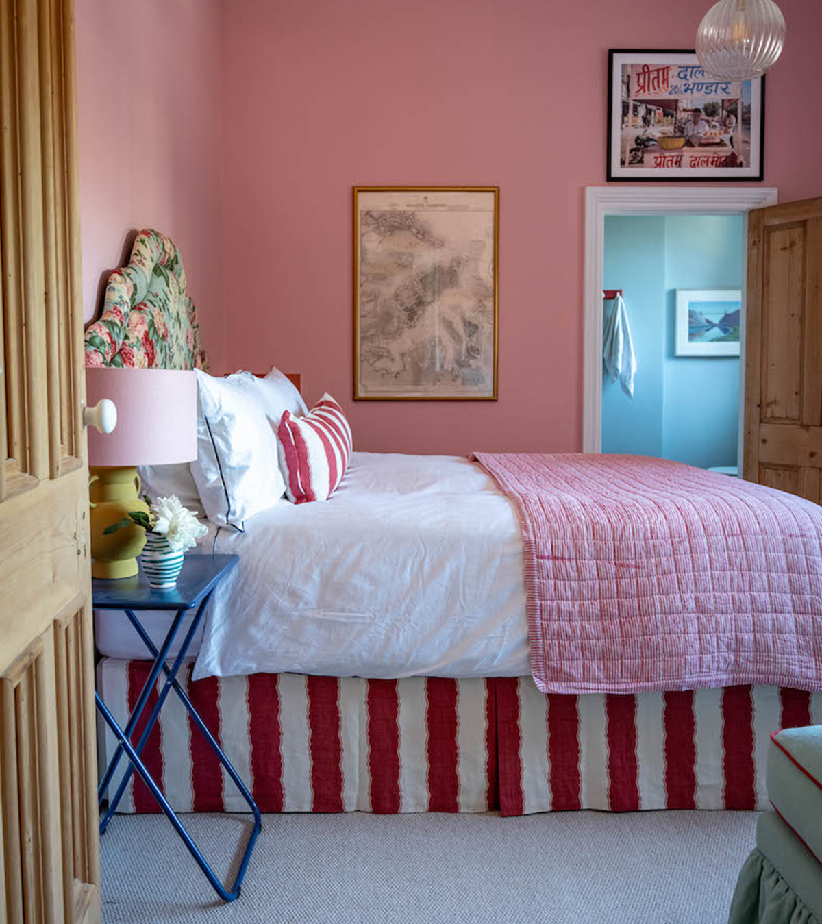 a pink bedroom with two pieces of art hanging on the wall, a double bed with a red and white stripe valance and a floral headboard and a blue metal bedside table with a yellow and pink table lamp