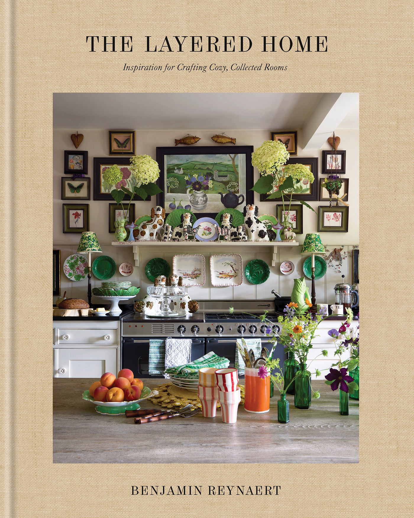 book cover for the layered home by Benjamin Reynaert depicting a kitchen filled with antique pieces such as staffordshire dogs and botanical artwork, in the foreground is a wooden island worktop with mini vases, striped tumblers and stacks of plates on top