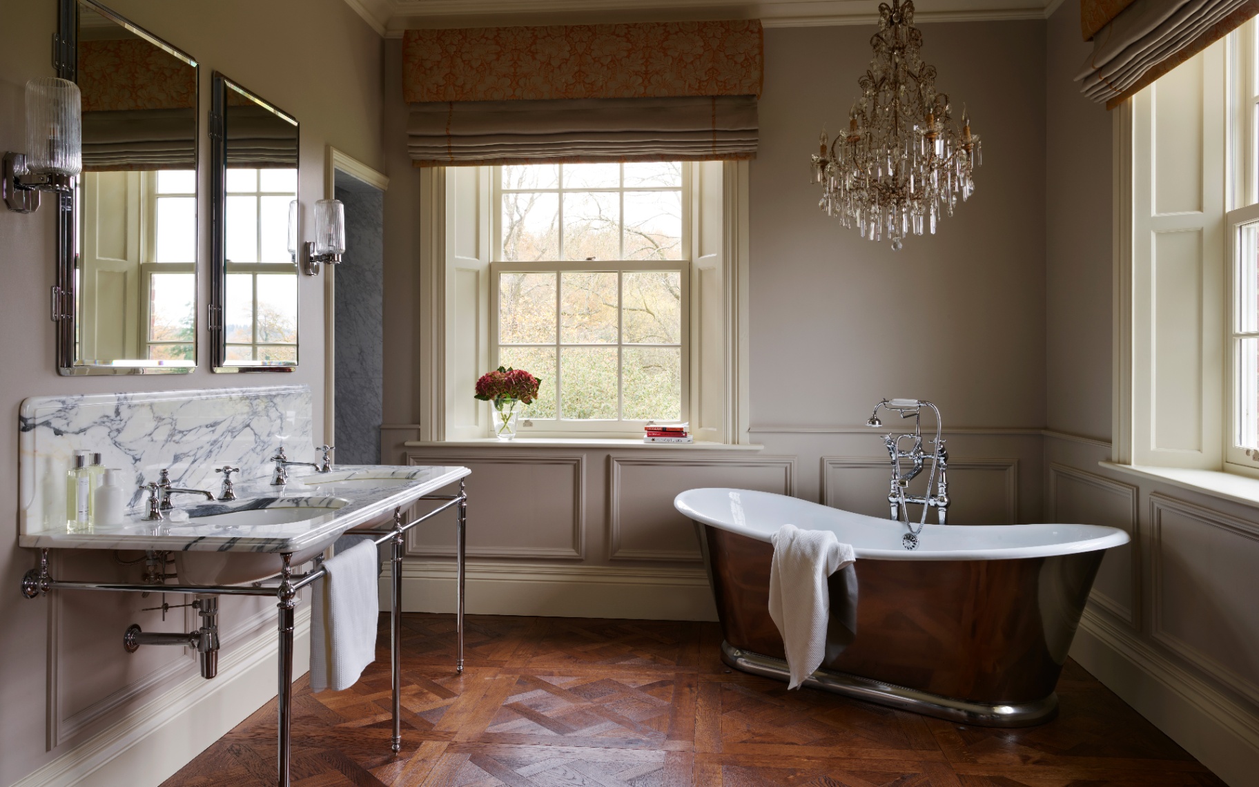 a traditional bathroom with a chrome freestanding bath, a large chandellier, pale brown panelled walls and a chrome pedestal sink