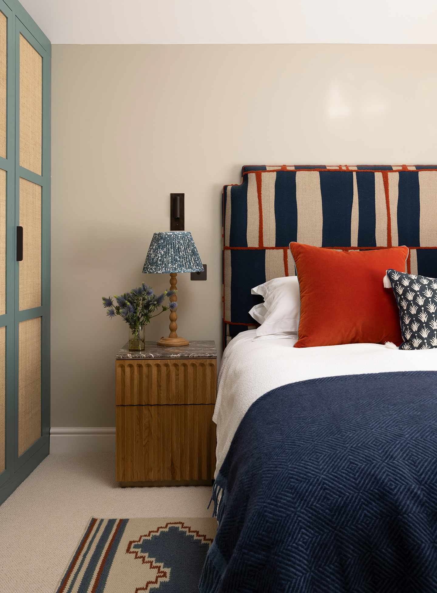 a close up shot of a bedroom with blue, cane-fronted wardrobes on the left and, on the right, a bed dressed with white linen, a navy blue throw and orange and blue cushions, with a headboard in a graphic fabric. A bedside table holds a lamp and flowers