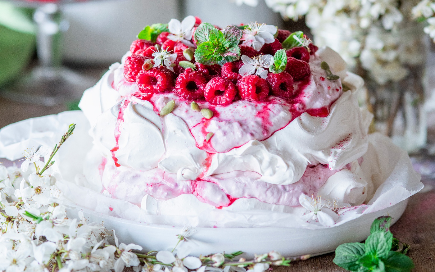 a white pavlova topped with a mound of fresh raspberries on a white dish and surrounded by fresh blossoms