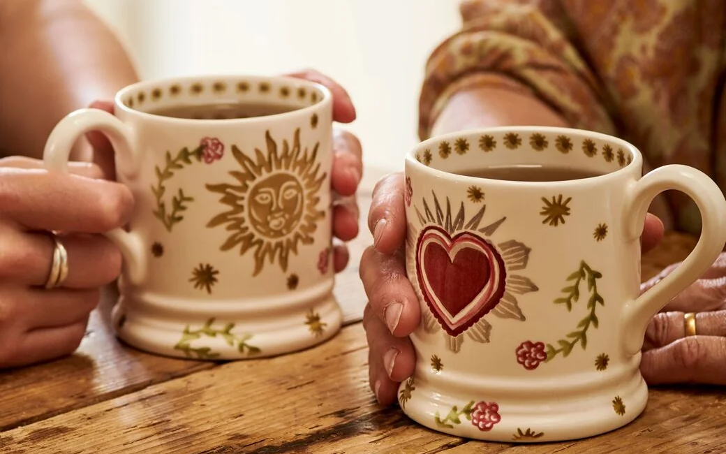 Emma Bridgewater Elizabeth collaboration pictured two mugs with designs of sun and heart being held by two people on a table 