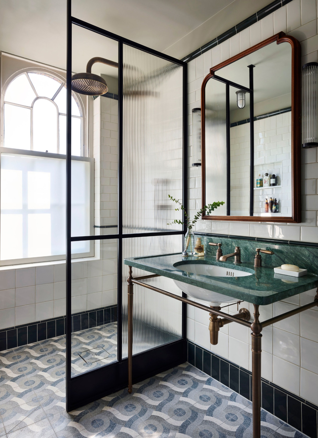a bathroom with a ribbed glass shower screen with black hardware, a large brass mirror, a pedestal sink with green marble top and a pale blue and white patterned tiled floor