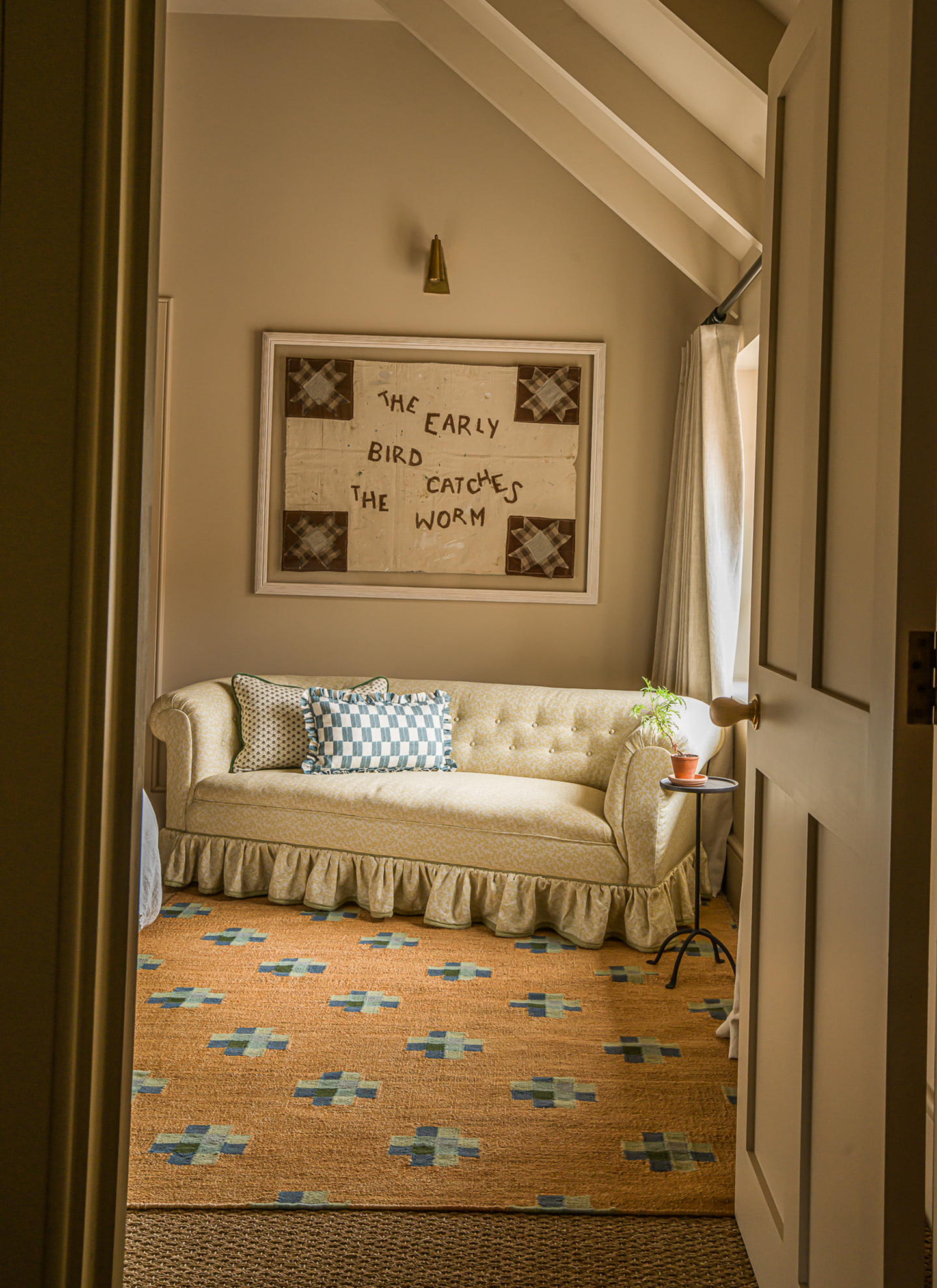 View through an open doorway into a cozy nook with a skirted cream daybed, checkered pillow, and patterned woven rug, topped with a framed quilt piece reading "The early bird catches the worm" on a neutral wall