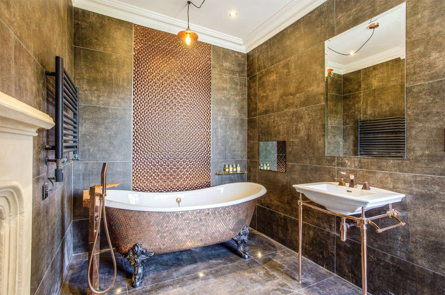a bathroom with brown marble floor and wall tiles, a copper bath and a pedestal sink