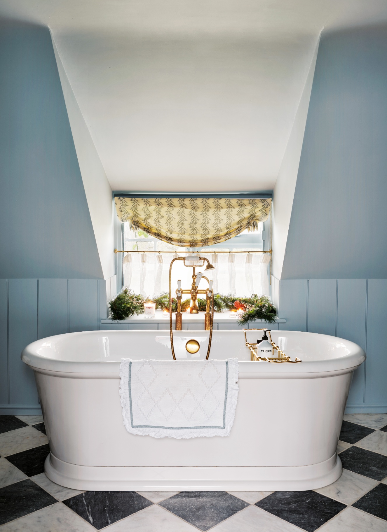 A bright bathroom with light blue wood panelling and a freestanding bath on checkerboard tiles. In the alcove window behind the brass fittings, a festive Christmas garland with warm lights rests along the deep sill
