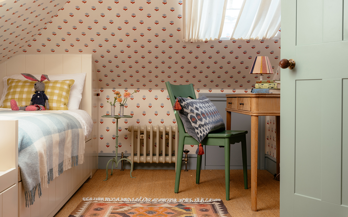 An attic bedroom with wallpapered walls and sloped ceiling. The skylight windows are dressed with light fabric curtains. A patterned rug sits on the floor, a bed to the left and a wood desk and green chair to the right