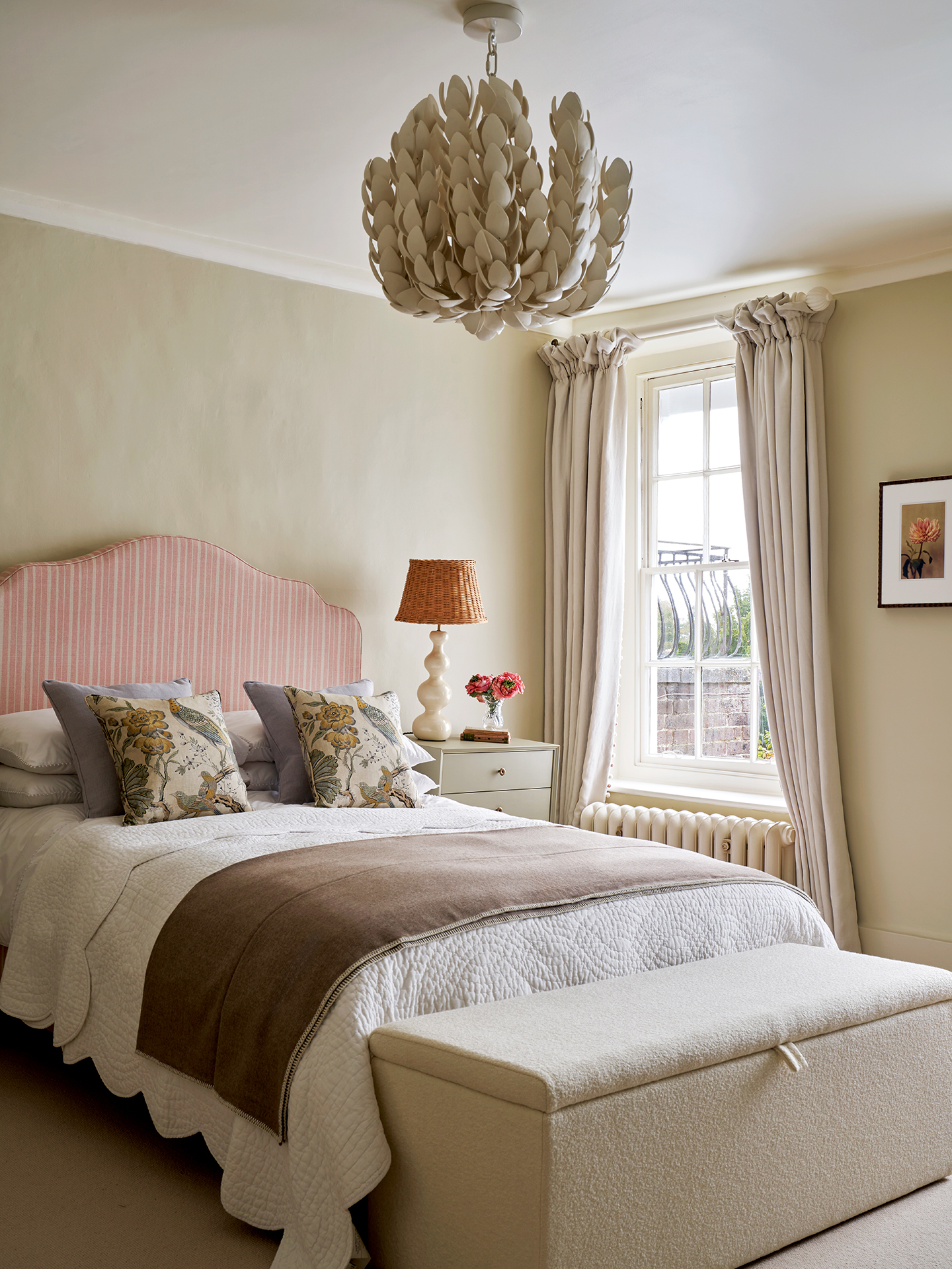 A neutral bedroom with a double bed, with a pink striped headboard, floral cushions and brown throw, neutral curtains, a bench at the end of the bed and neutral bedside tables topped with a table lamp and flowers. A sculptural leafy pendant light is above