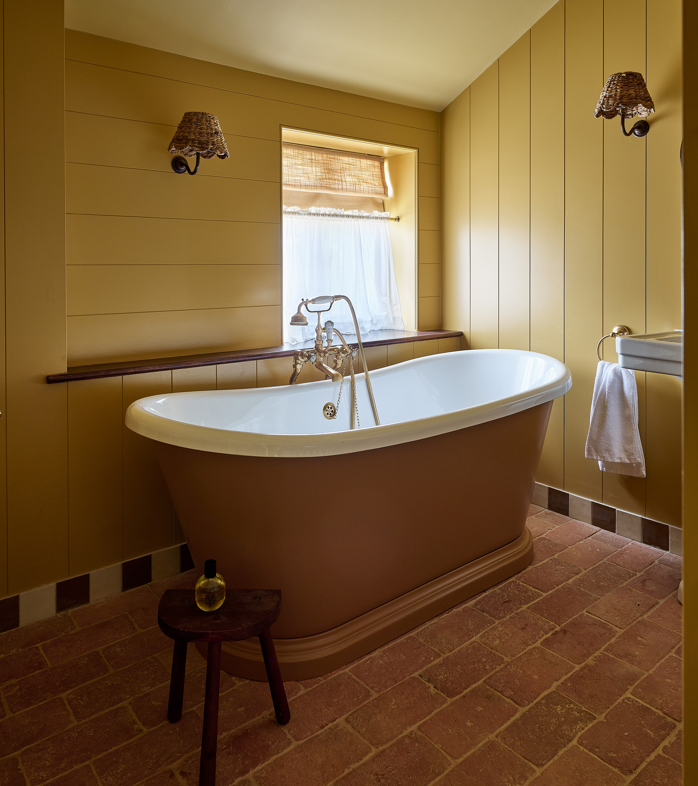 a bathroom with yellow panelled walls, flagstone flooring, a freestanding bath tub in mustard yellow and two wicker sconces