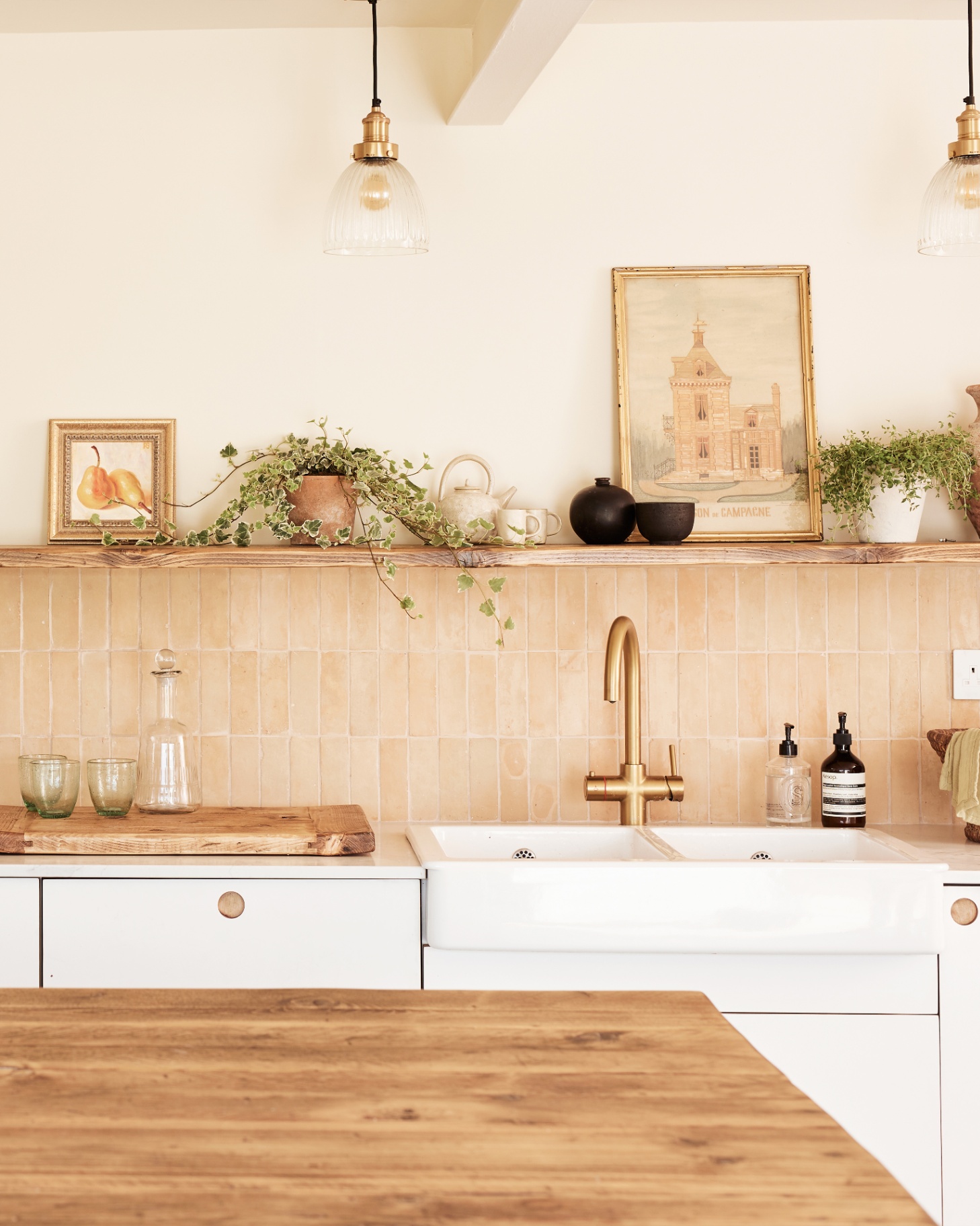 a kitchen sink with neutral zellige tiles on the wall, white cabinets and a long open shelf with plants and art