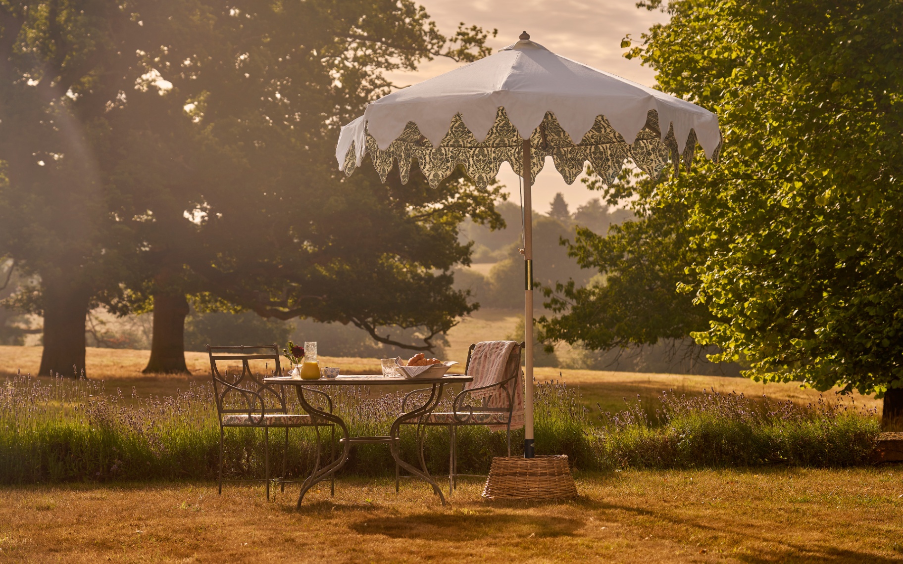 a cream parasol with a patterned canopy shading an iron bistro set in a field with long grass and trees behind it