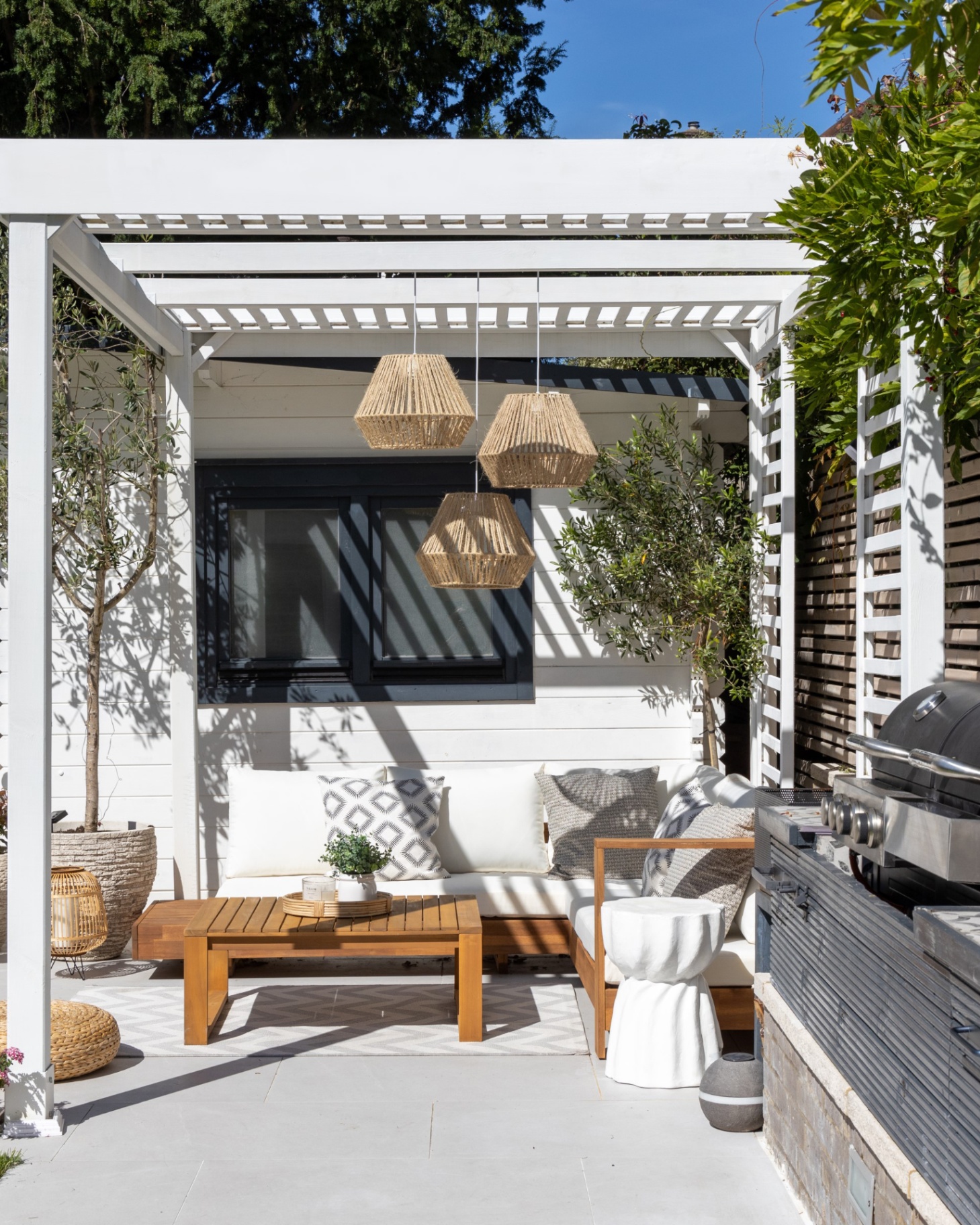 an outdoor kitchen area with a white pergola over a seating area with a wooden outdoor sofa with white cushions, rattan pendants hanging from the top and a bbq and kitchen area to the right