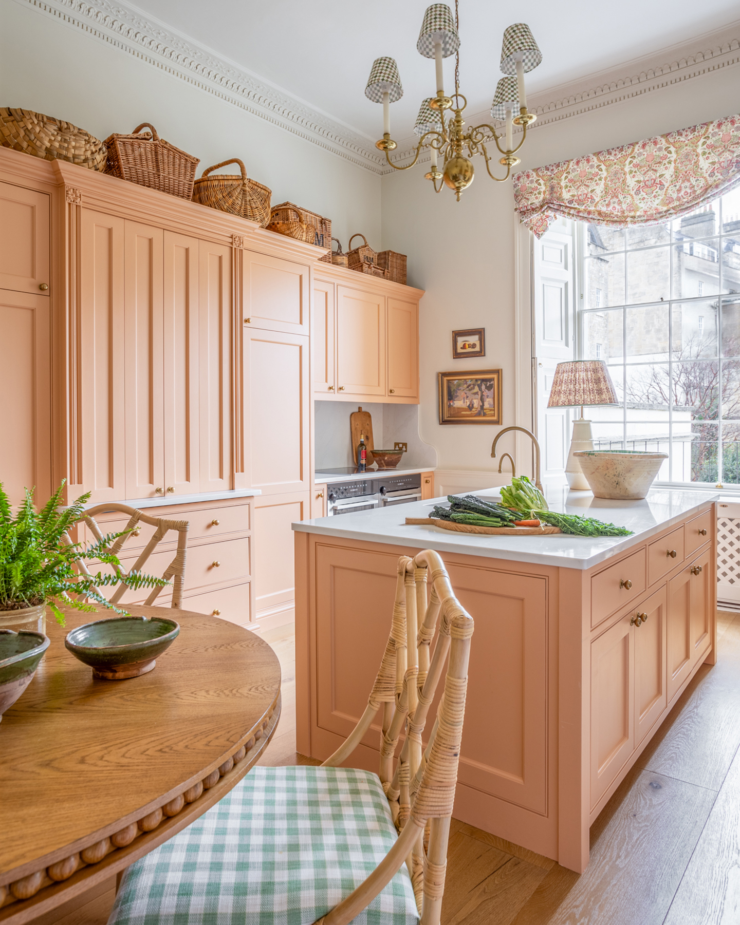 a kitchen with peach pink cabinets, a round wooden table with rattan chairs and green and white gingham seat pads, a chandelier with gingham lampshades and wicker baskets on top of the cabinets