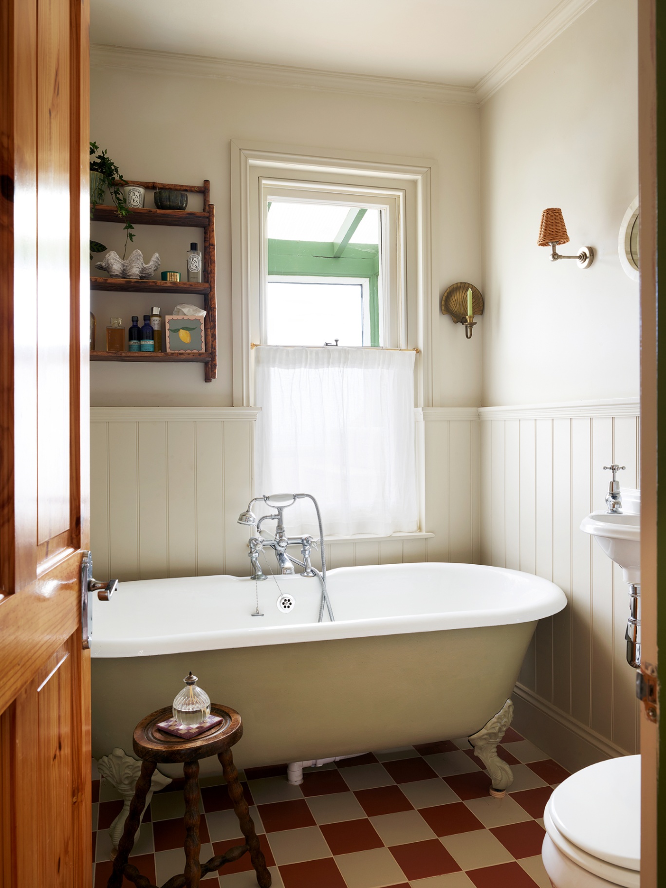 A bathroom with a checkerboard tiled floor, freestanding bath, wooden bobbin stool and tongue-and-groove panelling on the lower half of the walls. A cafe curtain covers the window and on the wall is a shelf with trinkets, candle sconce and wall light