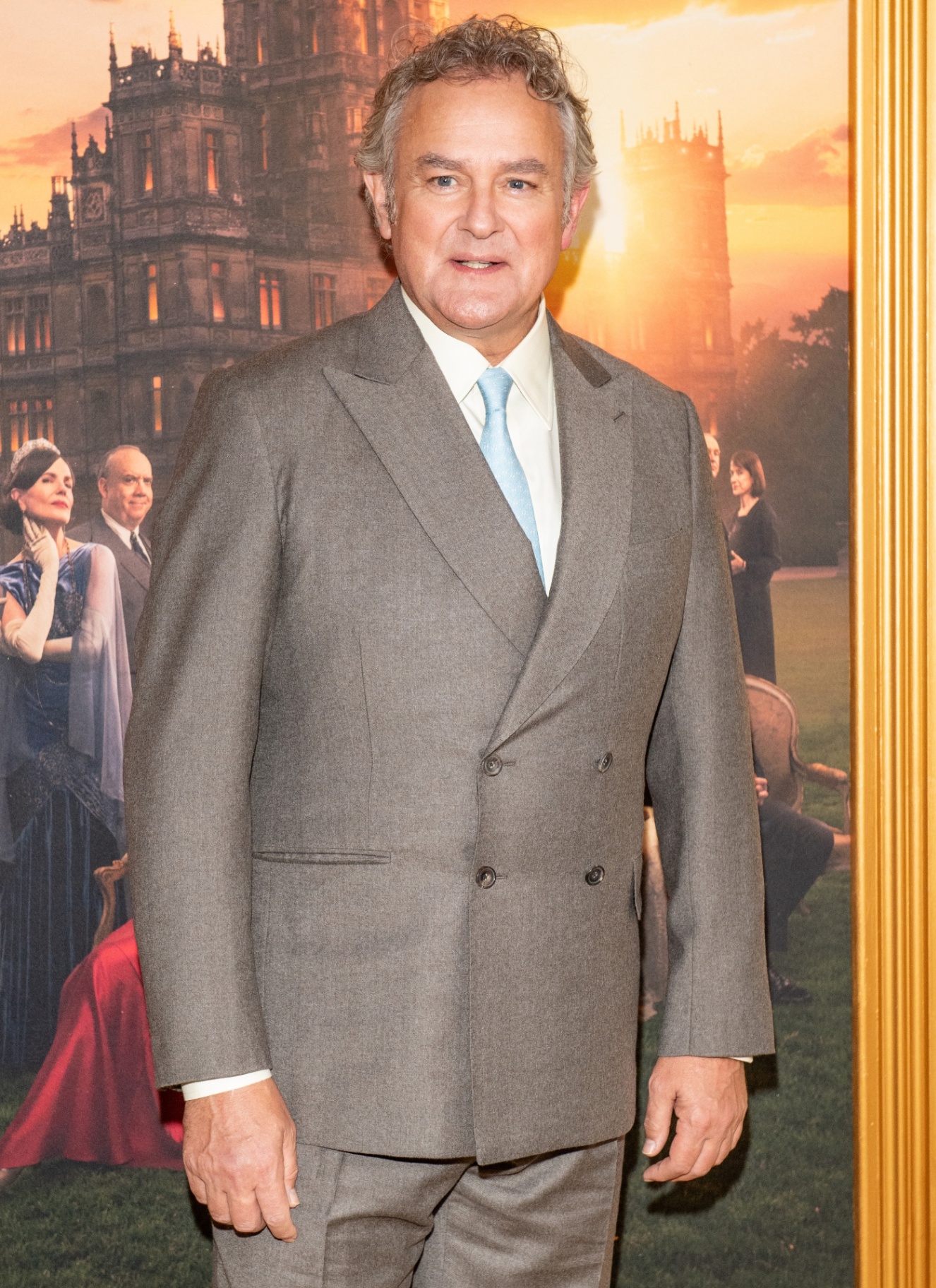 hugh bonneville wearing a grey suit and a blue tie on the red carpet promoting the downton abbey movie with a downton abbey poster background