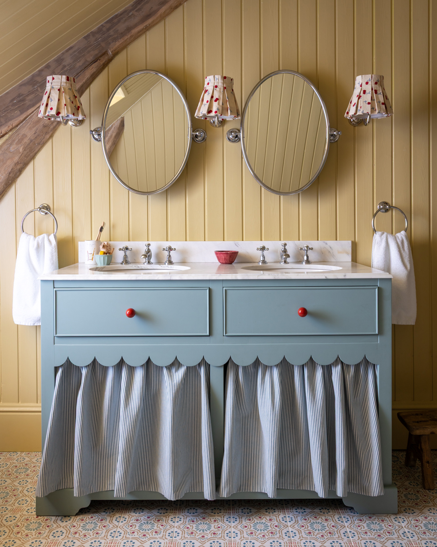 a bathroom with a pale yellow panelled wall, a pale blue painted double vanity with a stripe skirt, two oval mirrors above it wtih a set of 2 sconces either side of the mirrors