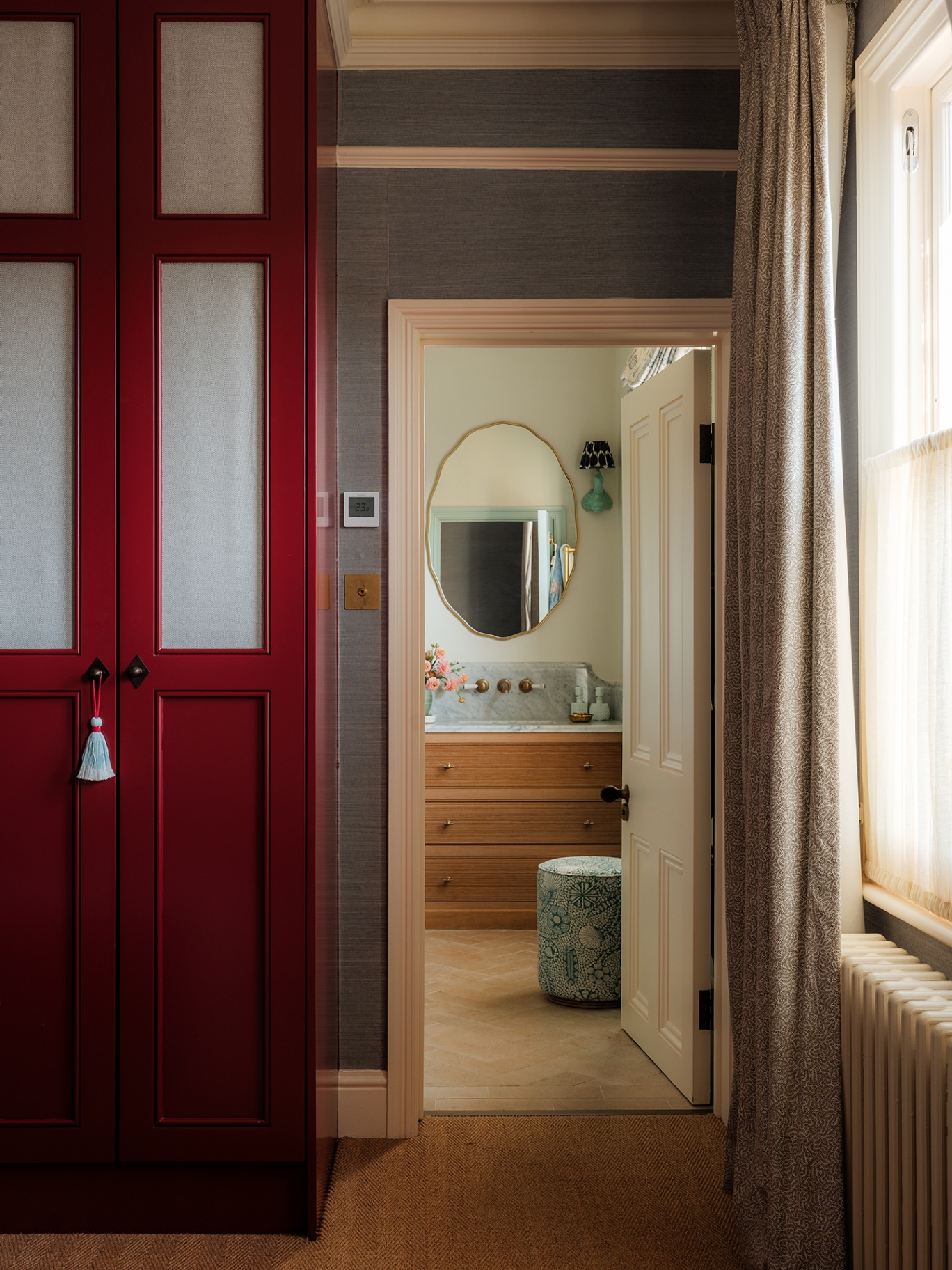 A view from a bedroom through to a bathroom with a large vanity, stool and oval mirror. In the foreground is a built-in red wardrobe with glass panels dressed with fabric and a tassel hanging from the handle