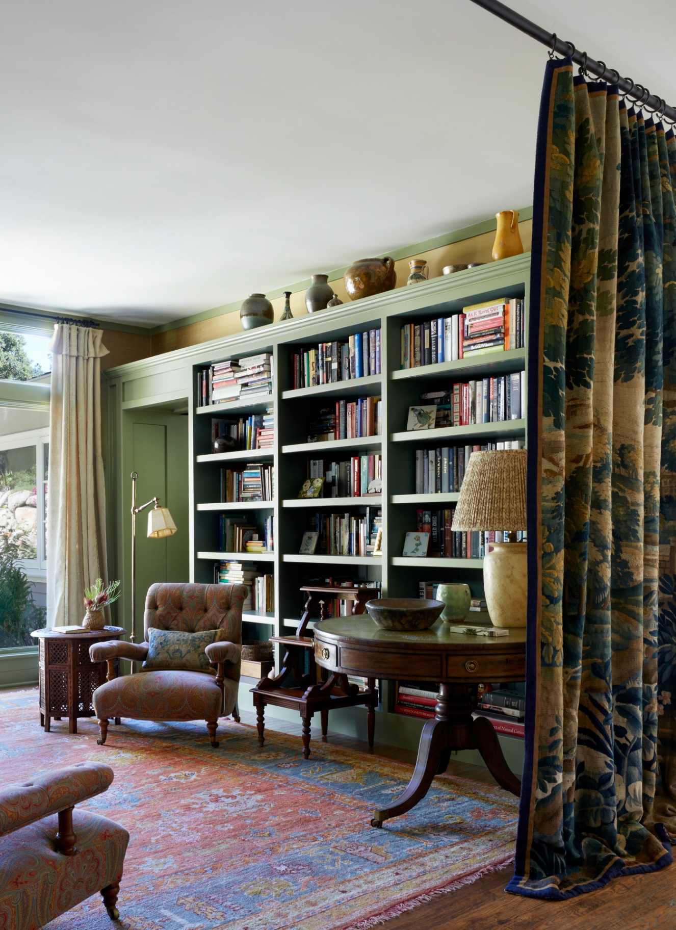 A secluded library reading space separated by heavy, floor-to-ceiling floral tapestry curtains, with green built-in bookcases and a classic armchair
